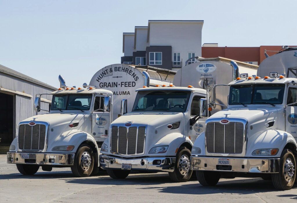 Three white tanker trucks at Hunt & Behrens Grain-Feed facility under daylight.